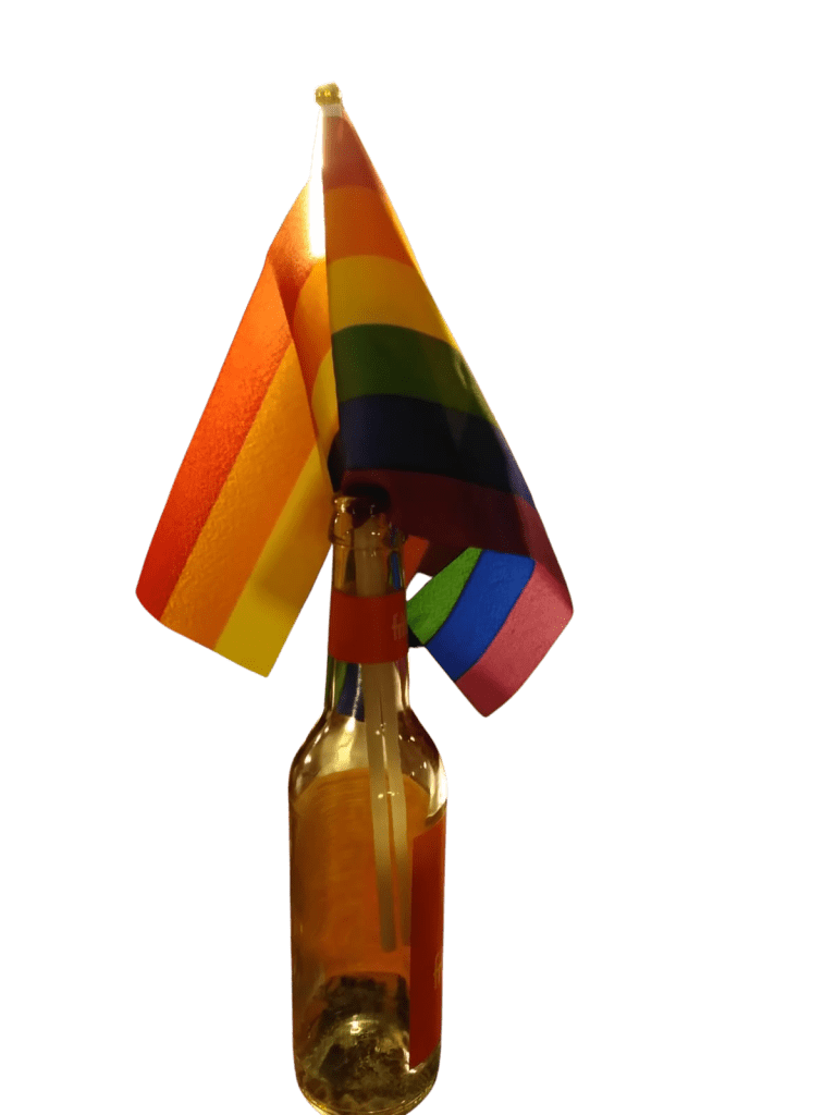Eine leere, braune Glasflasche, in der zwei kleine Regenbogenflaggen stecken.