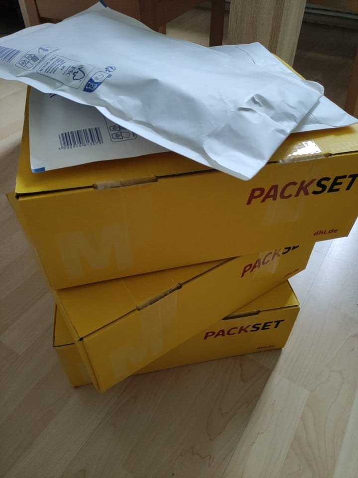 Foto von drei gelben Postpäckchen, die gestapelt auf einem hellen Holzboden stehen. Auf dem Stapel liegen außerdem zwei dicke weisse Umschläge.